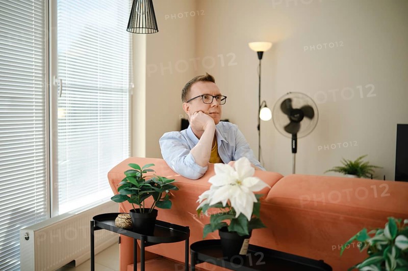 A man sits thoughtfully on a couch in a cozy living room, enjoying a moment of reflection. Bright natural light streams through large windows, illuminating indoor plants and stylish decor