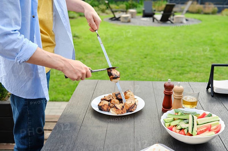 Unrecognizable man removes grilled meat from skewer to a plate. The concept of a picnic in the backyard