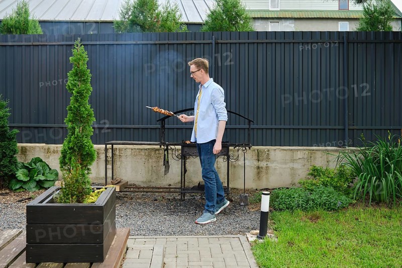 In a vibrant backyard garden, a man stands next to a grill, skillfully cooking skewered food. Lush greenery surrounds the outdoor space, creating a lively atmosphere during a sunny afternoon