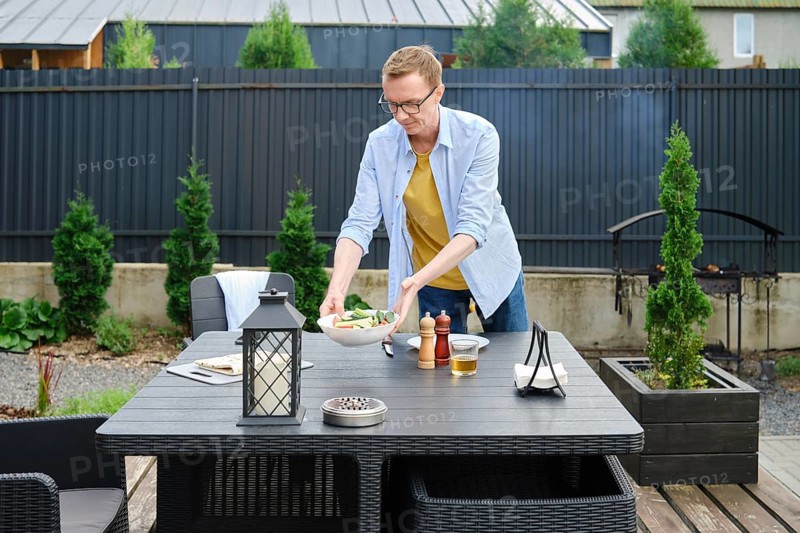 A man is arranging a fresh salad on a table in an inviting garden area. The sun is shining brightly, illuminating the surrounding greenery and modern outdoor furniture