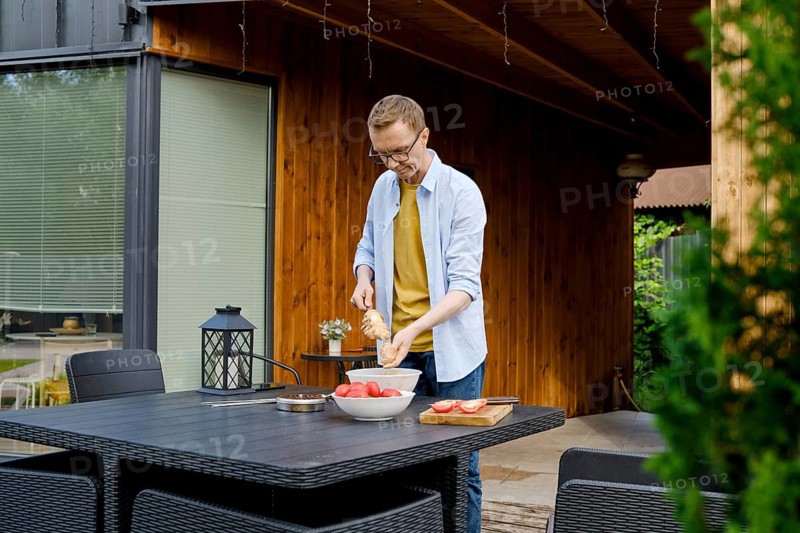 A man is joyfully preparing a meal outdoors at a wooden kitchen area. He strands meat on a skewer enjoying the warm afternoon sunlight and inviting environment