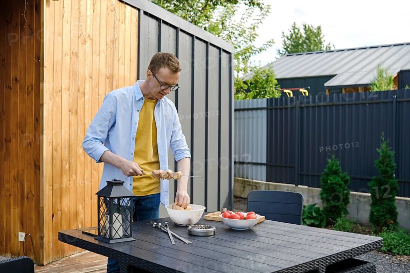 A man wearing a yellow shirt and denim jacket is preparing meat skewers at a wooden table in a backyard. Fresh vegetables are on the table, and greenery surrounds the area during a sunny day