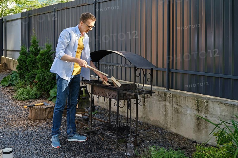 A middle-aged a man puts firewood in the grill in a backyard. He is going to cook meat outdoors on a sunny day