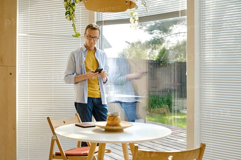 A middle-aged man stands in a bright, modern kitchen checking weather forecast at his smartphone. Sunlight filters through the blinds, illuminating the space