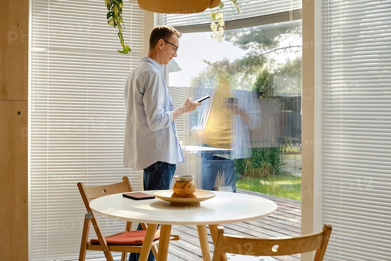 A man stands inside a stylish room, using his smartphone while looking out through a large glass window. Sunlight illuminates the space, and green plants are visible outside