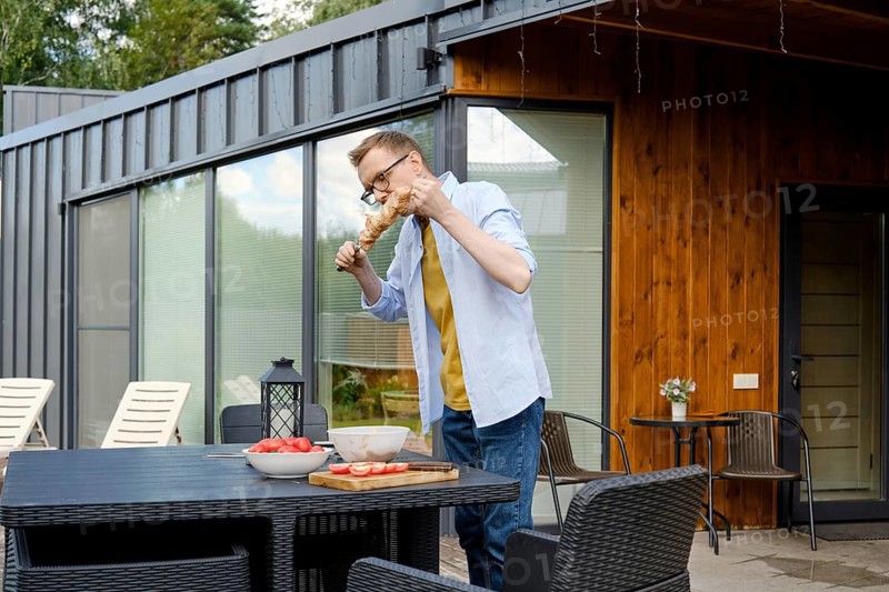 A man is enjoying a summer barbecue at a stylish wooden house. He is focused on grilling meat, surrounded by bowls of fresh ingredients and a pleasant outdoor setting
