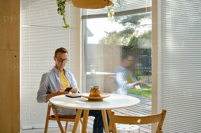 A middle-aged man in casual attire sits at a round table, focused on his smartphone. Sunlight streams through the blinds, illuminating the stylish indoor area
