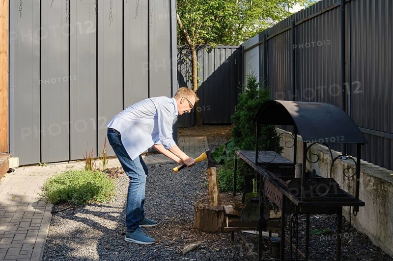 A middle-aged man is chopping a piece of wood in a well-maintained backyard garden. He is dressed casually in a light blue shirt and jeans while surrounded by greenery and a gray fence