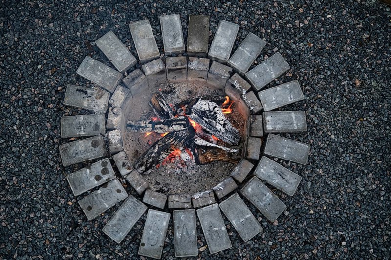 Bright flames dance among burning logs in a circular stone fire pit during early evening. The warm glow reflects off the dark gravel surrounding the fire, creating a cozy atmosphere under twilight