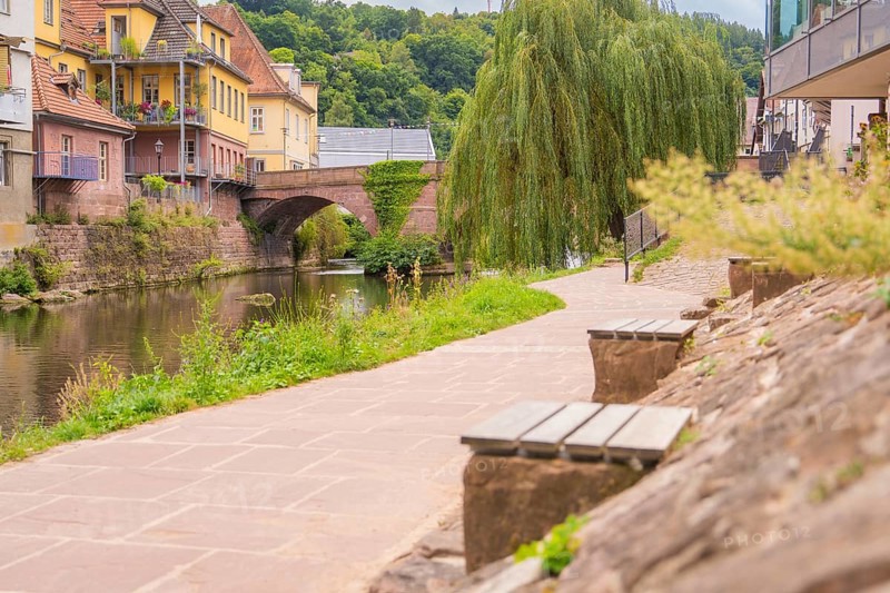 Paved path along a river with stone wall and benches, a bridge in the background, small town of Perle Calw, Black Forest, Germany