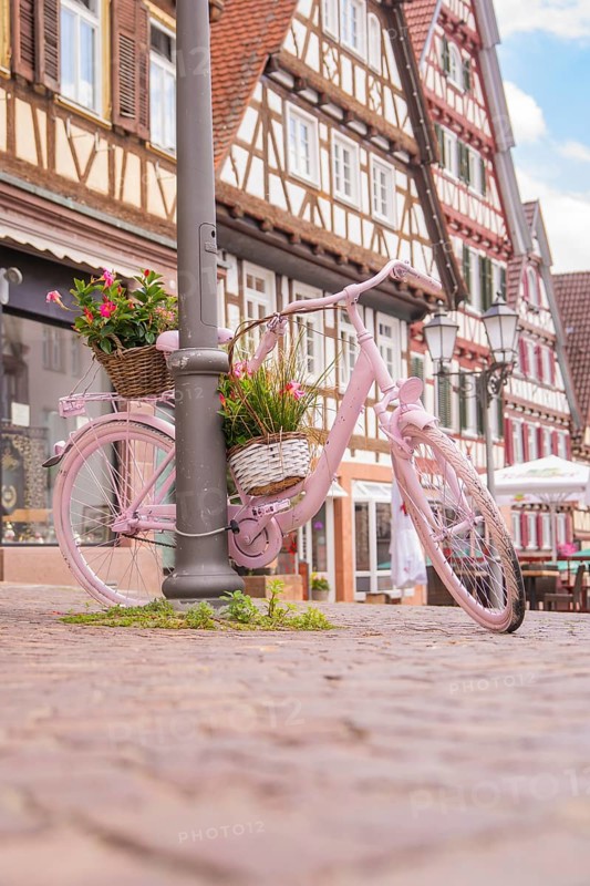 A pink bicycle with a basket of flowers in front of half-timbered houses on a cobbled path, small town of Perle Calw, Black Forest, Germany