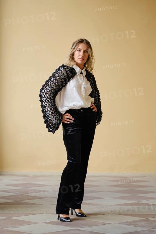 A middle-aged woman stands with hands on her hips in a trendy outfit featuring a unique sweater and sleek trousers. The neutral background highlights her chic look and modern style