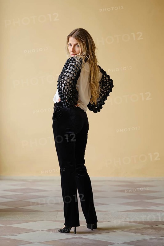 A middle-aged woman stands confidently with her back turned, showcasing a chic black and white outfit. The setting features a clean, light-colored wall and patterned tiles that enhance her fashionable presence