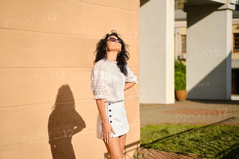 A woman stands playfully against a tan wall in a bright outdoor environment, enjoying the warm sun. She wears a white blouse and mini skirt and facing the sun