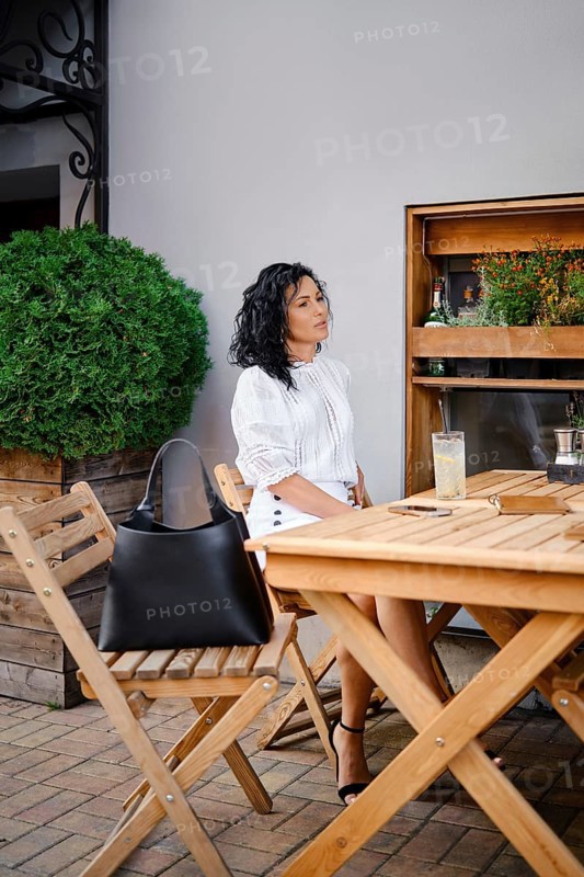A woman with curly hair relaxes at a wooden table in an outdoor cafe, dressed in a fashionable white blouse. She enjoys a cold drink under the warm sun, surrounded by greenery and stylish decor