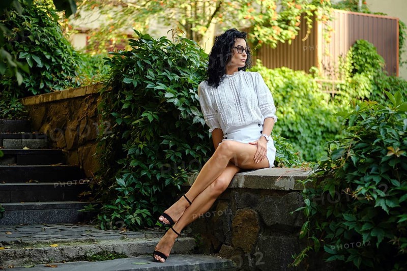 A woman with curly dark hair and glasses sits on a stone wall in a garden, surrounded by greenery. She wears a stylish outfit and exudes confidence while enjoying the peaceful atmosphere