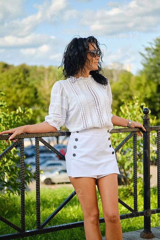 In a vibrant park setting, a woman poses confidently in a white blouse and mini skirt. Sunlight filters through trees, highlighting her relaxed demeanor and chic fashion choice on a beautiful day