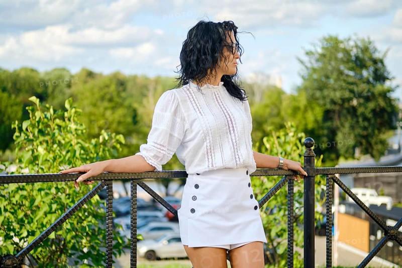 A woman with curly hair stands on a balcony, leaning against a railing. She wears a white blouse and a white skirt. The setting is bright and sunny with lush greenery in the background
