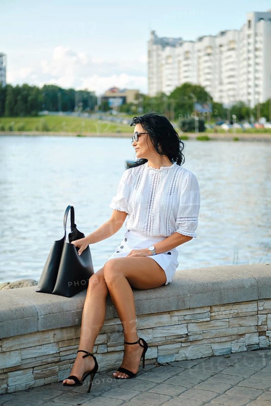 A fashionable woman sits by the water, dressed in a white top and shorts while wearing stylish heels. She gazes towards the cityscape, exuding confidence against a sunny backdrop