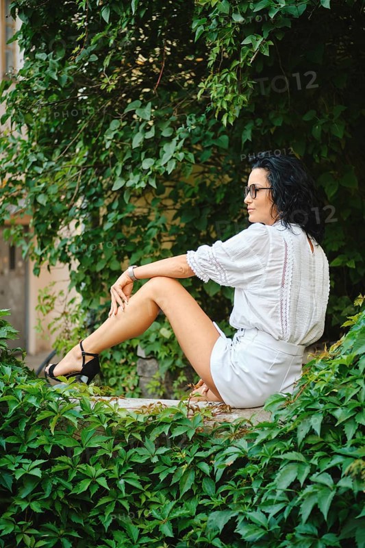 A woman with dark hair sits peacefully in an urban garden surrounded by vibrant green leaves. She wears a white outfit and sunglasses, exuding a relaxed vibe on a sunny day