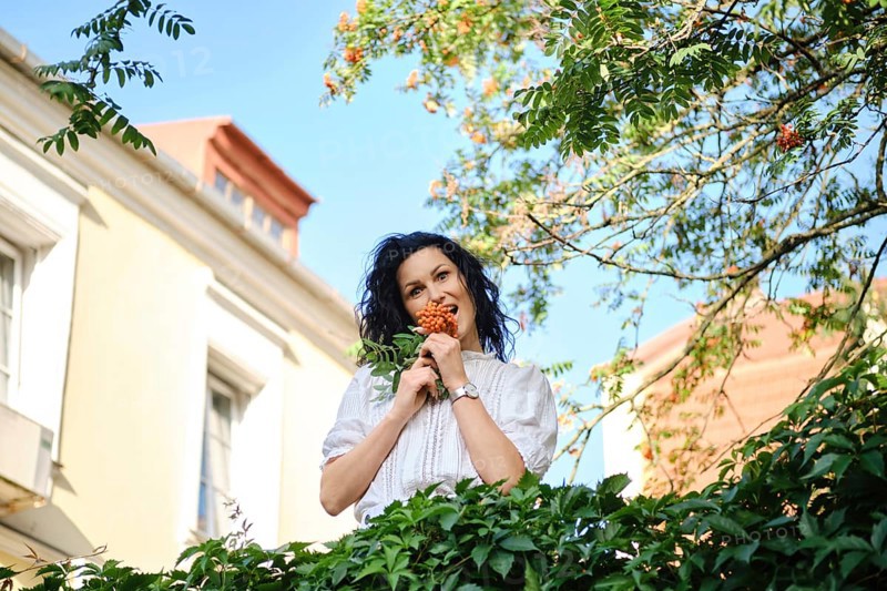 A playful woman stands surrounded by greenery holding a cluster of bright berries. The sun shines down, highlighting her joyful expression as she enjoys the beauty of the garden setting