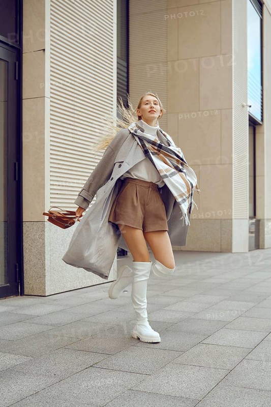 A model captures attention on a bustling city sidewalk, showcasing a trendy ensemble featuring a long coat, a cozy scarf, and fashionable knee-high boots