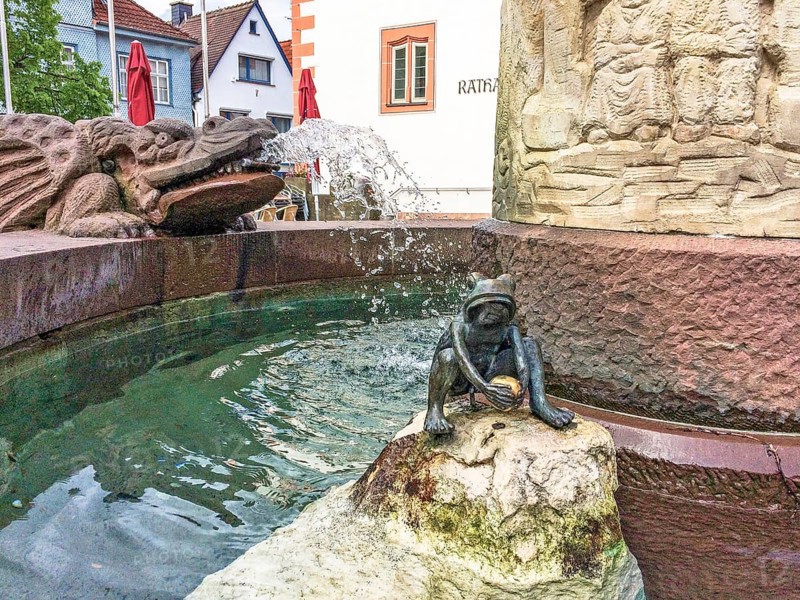 Sculpture figure of a frog holding a golden ball depicting the Frog King from the Brothers Grimm fairy tale in a fairy tale fountain, gargoyle in the form of a winged dragon behind it on the left, Steinau an der Straße, Hesse, Germany
