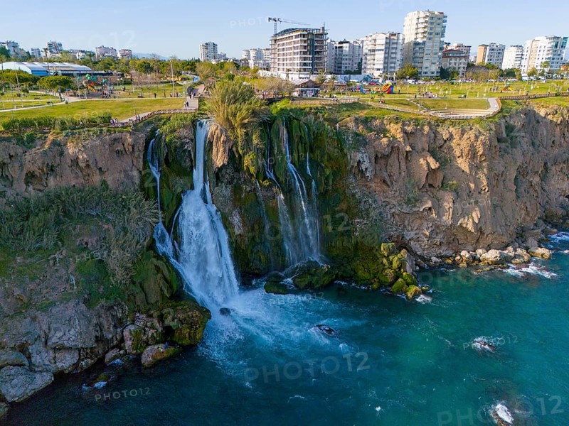 A waterfall cascades from a cliff into the sea, surrounded by urban landscape and green parks, aerial view, Lower Düden Waterfall, Düden River, Antalya, Turkey