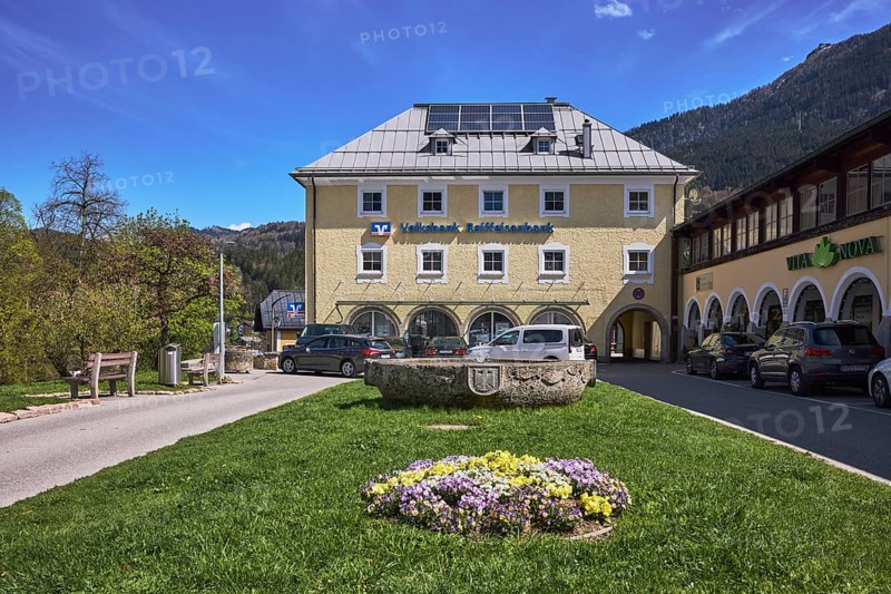 Volksbank Raiffeisenbank Oberbayern Südost eG, Fischerbrunnen, master stonemason August Wolf, fountain, building, car park, vehicles, lawn, flower bed, trees, mountain landscape, mountains, coniferous forest, blue sky, cirrostratus clouds, Am Fischerbichl, Bahnhofstraße, Berchtesgaden, Upper Bavaria, Berchtesgadener Land district, Bavaria, Germany