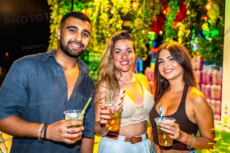 Three friends smiling and holding cocktails at a vibrant outdoor night party, enjoying the festive atmosphere