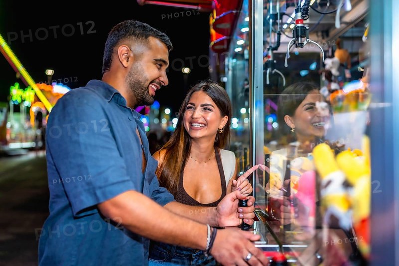 Young couple enjoying playful moments while trying to grab a prize from a brightly lit claw machine at a lively night carnival