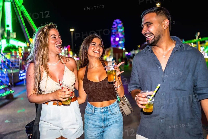Three joyful friends walking and laughing together at an amusement park at night, enjoying colorful cocktails under vibrant lights