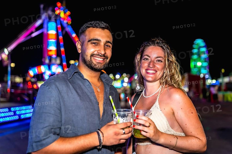 Young man and woman smiling, holding cocktails, enjoying a night out at amusement park with colorful lights in background
