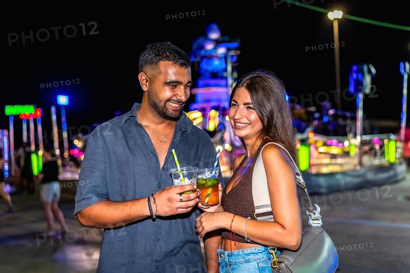 Young couple smiling and holding colorful cocktails at a lively funfair, celebrating nightlife and enjoying joyful moments together