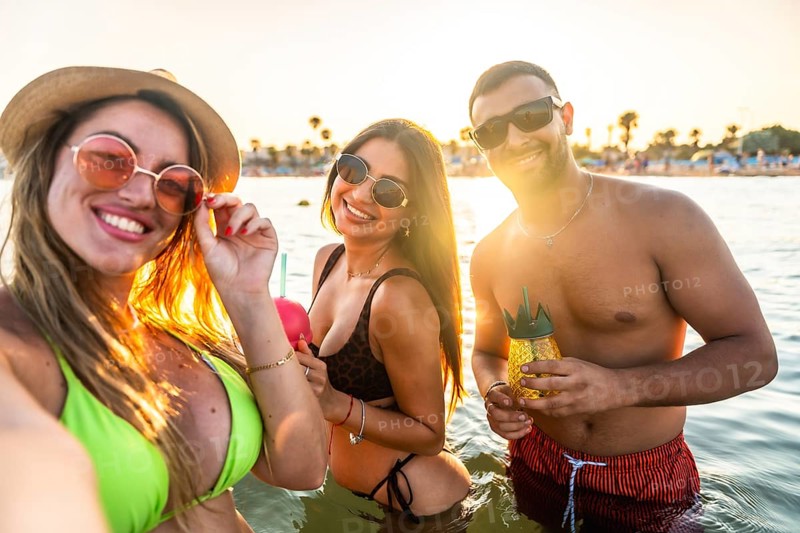 Young men and women having fun in the sea taking a selfie at sunset, enjoying refreshing cocktails