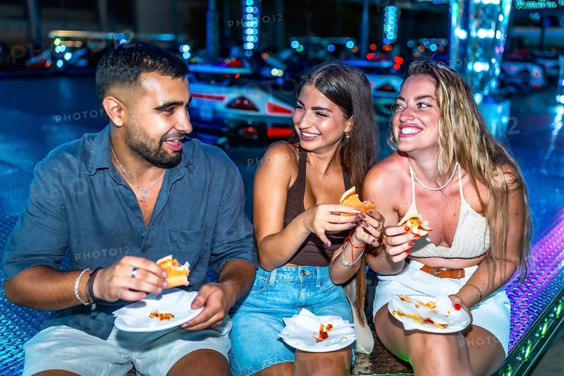 Three happy friends are enjoying burgers and pizza at an amusement park, illuminated by colorful lights at night