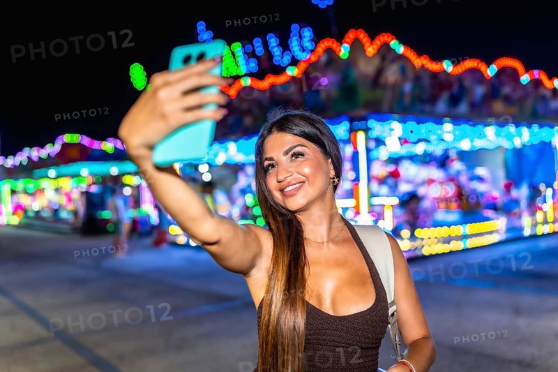 Happy woman taking a selfie at amusement park with her smartphone at night, colorful lights in the background