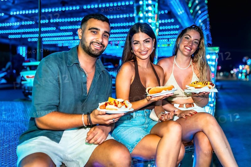 Three friends are sitting and smiling, holding hot dogs on paper plates at a brightly lit amusement park at night