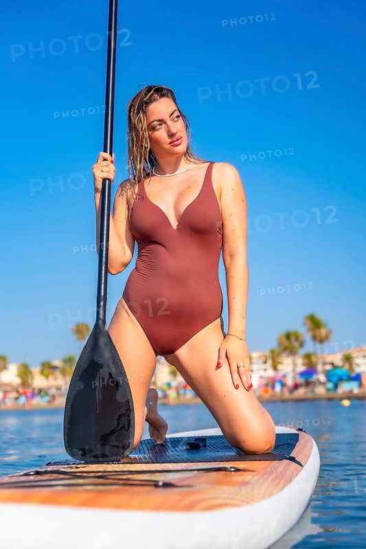 Young woman kneeling on paddle board, holding paddle and looking away, enjoying summer sunny day at the beach