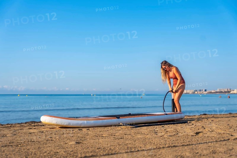 Woman inflating a paddleboard on a sandy beach, ready for a summer day of paddle surfing under a clear blue sky