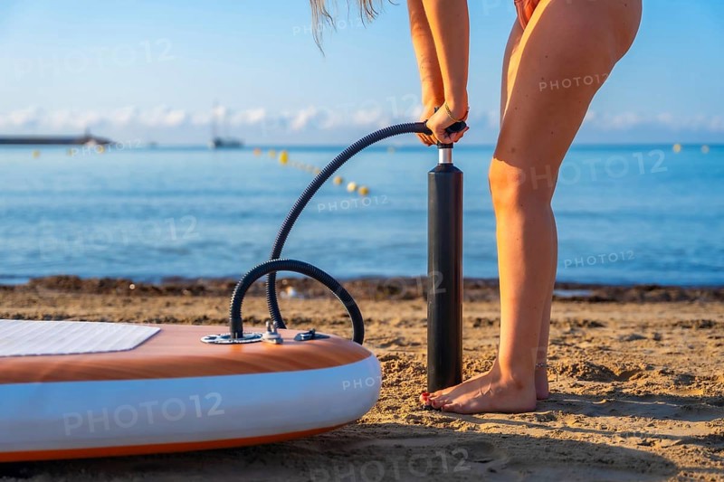 Woman inflating a paddleboard on a sandy beach, preparing for a summer paddle surfing session. Calm ocean and clear sky in the background