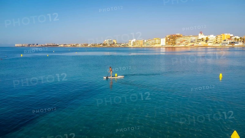 Calm waters and clear skies set the scene for a serene paddle boarding experience near a coastal town, highlighting summer leisure and tranquility