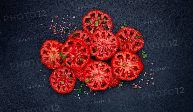 Freshly sliced ripe tomatoes arranged on a textured dark surface with herbs and salt scattered around
