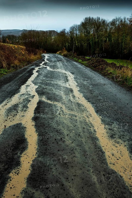 A winding rural road shows signs of heavy rain, with puddles forming along its surface. Puy de Dome. Auvergne Rhone Alpes. France