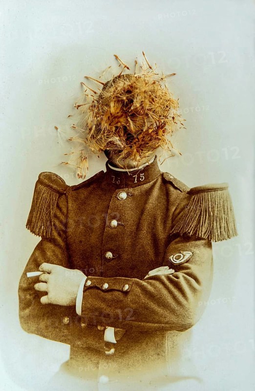 A soldier stands confidently, sporting a distinctive uniform with epaulettes, while a curious arrangement of flowers covers his head. The sepia tones highlight the unusual composition and moment