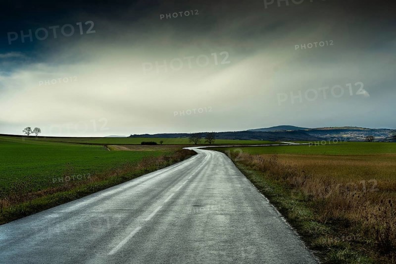 A winding road stretches through green fields under a cloudy sky, showcasing the beauty of the landscape with distant mountains in view. Auvergne. France