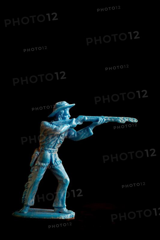 This vintage toy cowboy stands poised, aiming a rifle with intense focus. Dramatic lighting enhances its details against a stark, dark background