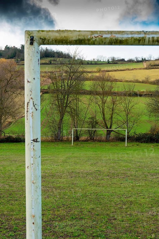 A weathered soccer goalpost stands in the foreground of a wide grassy field, with lush green hills and trees in the background, Auvergne. France