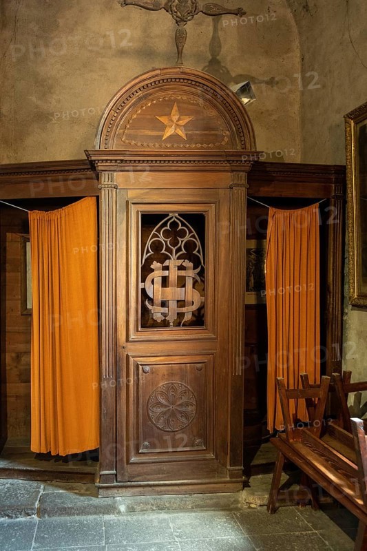 A beautifully crafted wooden confessional booth stands in a historic church, featuring ornate details and vibrant orange curtains. Auvergne. France