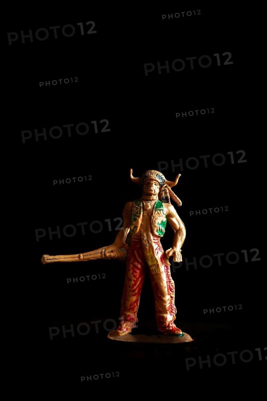 Vintage Native American Indian toy figure stands boldly in a dark setting, wielding a long weapon